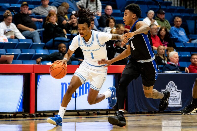Player in white uniform dribbles past black-uniformed defender as spectators watch from blue bleachers.
