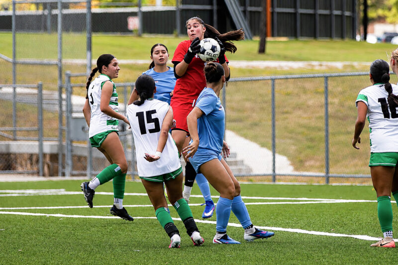 Soccer goalie in red jumps to catch the ball as players in green and blue crowd around her on the field.