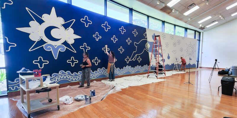 People paint a blue and white mural of stars, moon, and abstract shapes on a long wall in a bright room with art supplies.