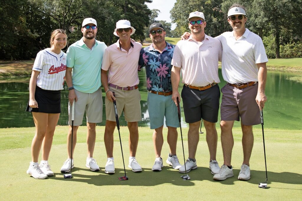 Six people on a sunny golf course green by a pond, holding clubs and smiling; five men in polos, one woman in a jersey and skirt.