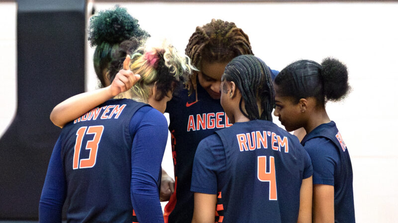 Four basketball players in navy RUNEM jerseys huddle on the court, heads bowed and arms around each other before the game.