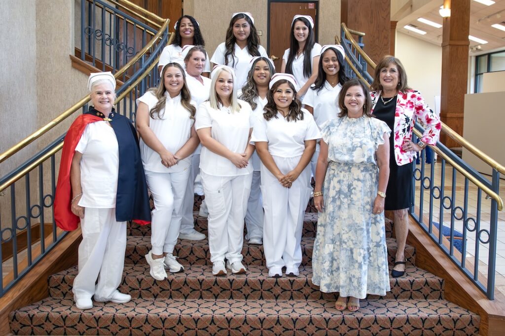 Women, mostly in white nursing uniforms, stand smiling on indoor stairs; two in floral dresses and one in a navy cape are at the front.