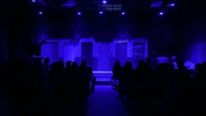 A blue-lit stage with multiple doors and backdrop faces seated audience silhouettes, just before a theater performance starts.
