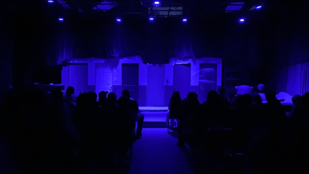 A blue-lit stage with multiple doors and backdrop faces seated audience silhouettes, just before a theater performance starts.