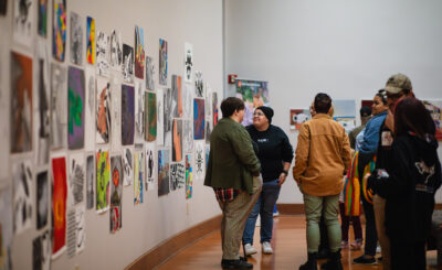 People converse in a gallery with colorful art on white walls, wooden floors, and a relaxed, social atmosphere.