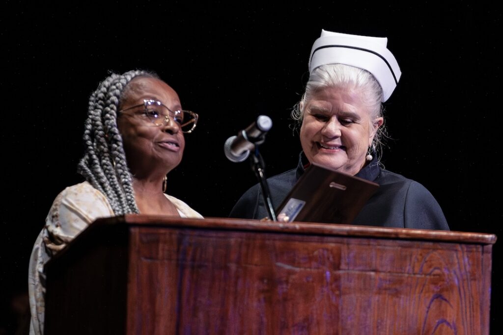 Two women at a podium face the audience; one speaks into a microphone, the other in a white nurse’s cap smiles, holding a plaque.
