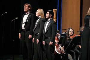 Three singers in tuxedos perform on stage with microphones, backed by string musicians and a visible cellist in warm concert hall lighting.