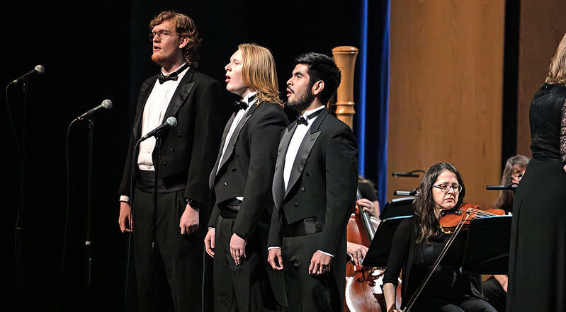 Three singers in tuxedos perform on stage with microphones, backed by string musicians and a visible cellist in warm concert hall lighting.