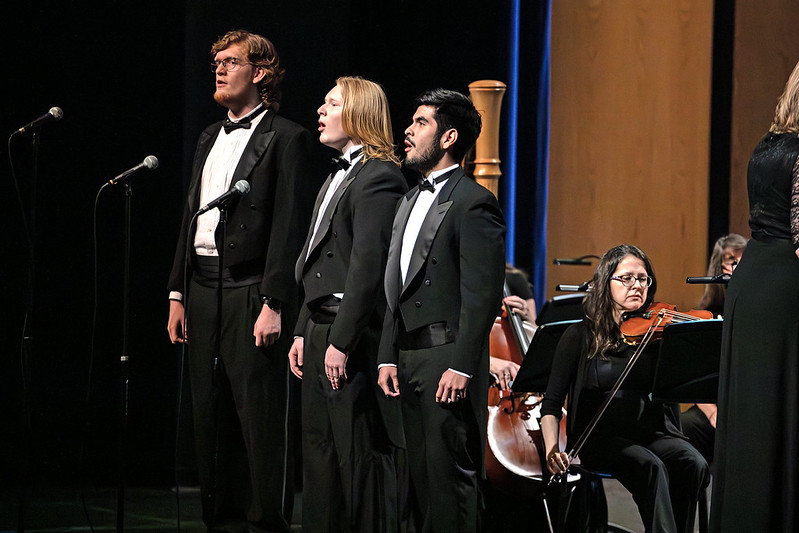 Three singers in tuxedos perform on stage with microphones, backed by string musicians and a visible cellist in warm concert hall lighting.
