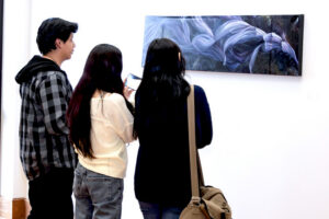 Three people stand before a rectangular painting of flowing purple and white forms; one takes a photo with their phone.