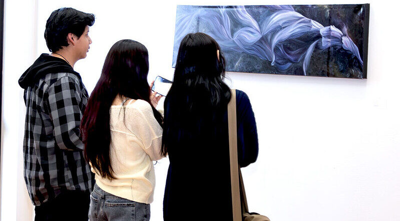 Three people stand before a rectangular painting of flowing purple and white forms; one takes a photo with their phone.