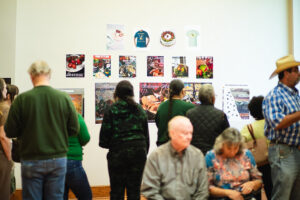 Visitors observe a wall display of colorful posters and t-shirts; some stand, others sit on nearby chairs.