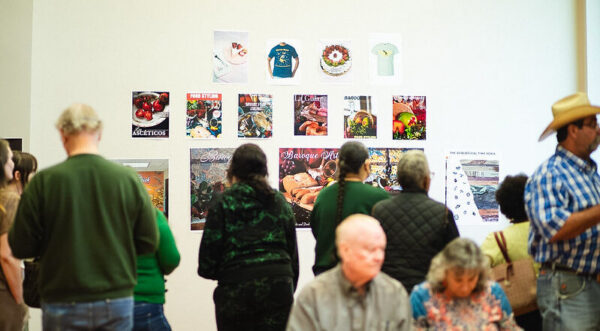 Visitors observe a wall display of colorful posters and t-shirts; some stand, others sit on nearby chairs.