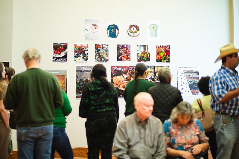 Visitors observe a wall display of colorful posters and t-shirts; some stand, others sit on nearby chairs.