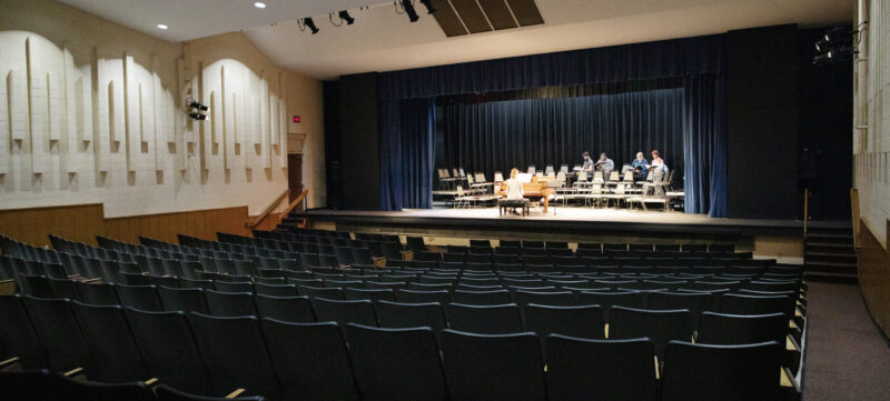 Rows of empty black seats face a well-lit stage with people seated, rehearsing, and a blue curtain backdrop.