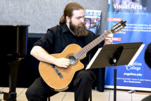 Man with long hair and a beard plays classical guitar by a piano, reading sheet music. Angelina College banners in the background.