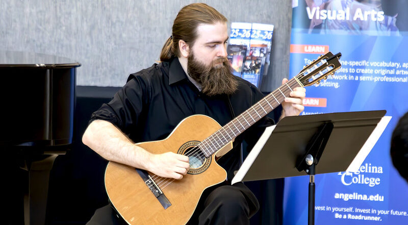 Man with long hair and a beard plays classical guitar by a piano, reading sheet music. Angelina College banners in the background.