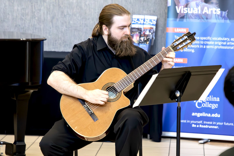 Man with long hair and a beard plays classical guitar by a piano, reading sheet music. Angelina College banners in the background.