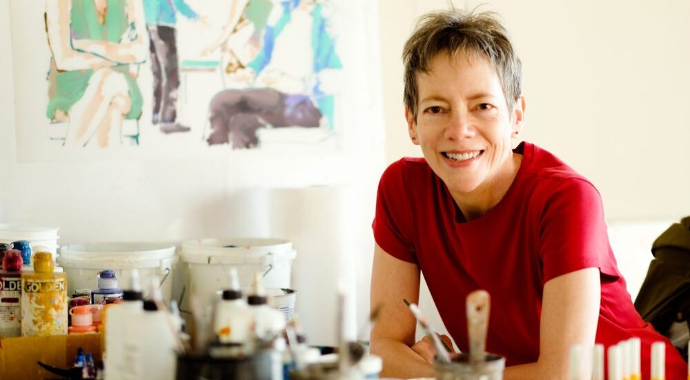 Person with short hair in a red shirt smiles at a table with art supplies; colorful painting in the background.