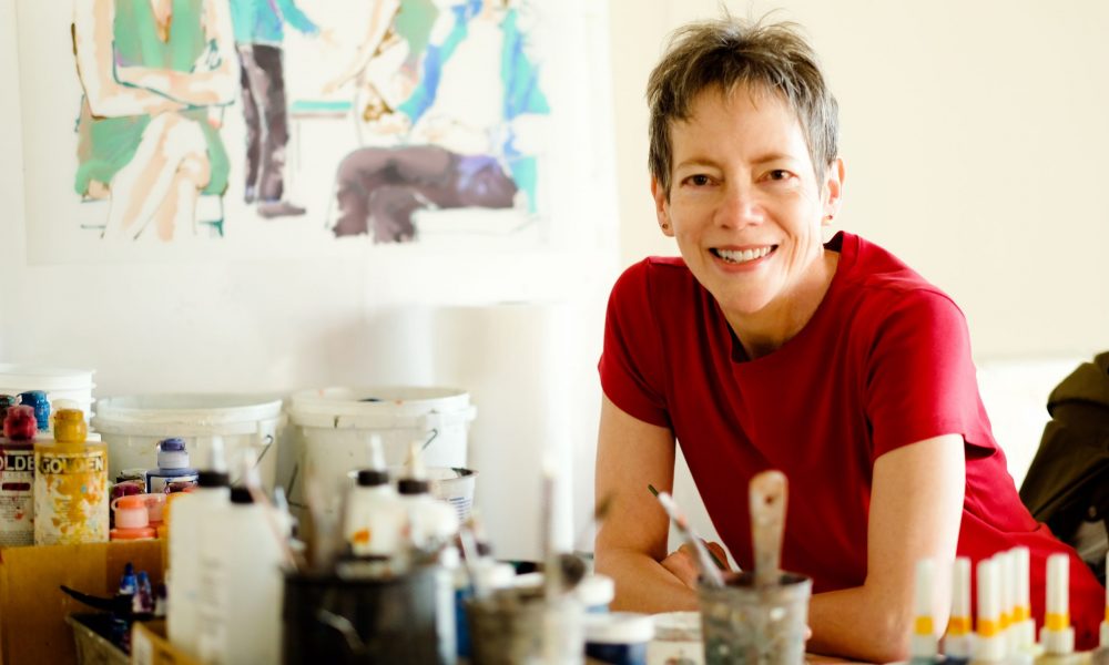 Person with short hair in a red shirt smiles at a table with art supplies; colorful painting in the background.