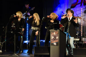 Four singers in black with silver accents perform on stage; guitarist, drummer, stage lights, and The Hit Parade sign in background.