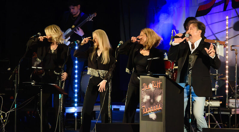 Four singers in black with silver accents perform on stage; guitarist, drummer, stage lights, and The Hit Parade sign in background.