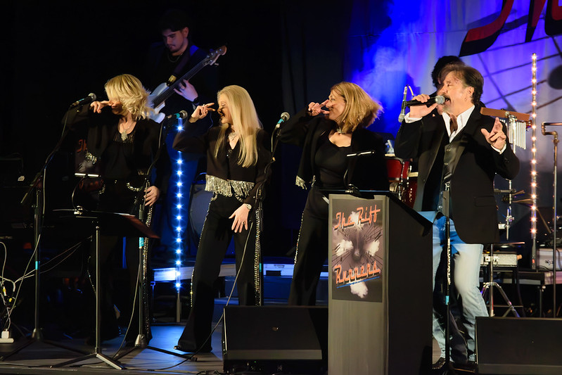 Four singers in black with silver accents perform on stage; guitarist, drummer, stage lights, and The Hit Parade sign in background.