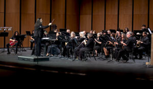 Conductor leads a seated orchestra on stage; musicians in black play woodwinds, brass, and percussion before a wooden backdrop.