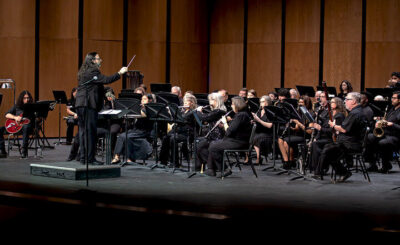 Conductor leads a seated orchestra on stage; musicians in black play woodwinds, brass, and percussion before a wooden backdrop.