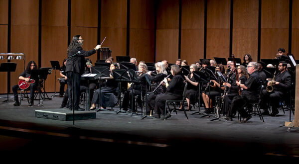Conductor leads a seated orchestra on stage; musicians in black play woodwinds, brass, and percussion before a wooden backdrop.