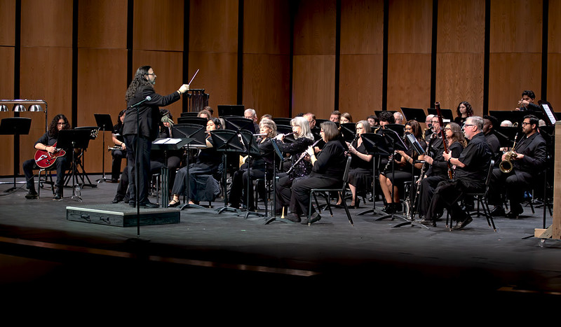 Conductor leads a seated orchestra on stage; musicians in black play woodwinds, brass, and percussion before a wooden backdrop.