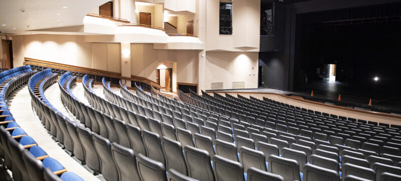 Curved rows of empty blue and gray seats face a stage with black curtains in a bright theater; balcony and beige walls behind.