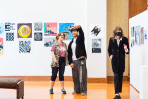 Three people in an art gallery with colorful abstract paintings; two smile for a selfie as a masked person in a coat walks by.