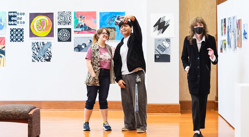 Three people in an art gallery with colorful abstract paintings; two smile for a selfie as a masked person in a coat walks by.