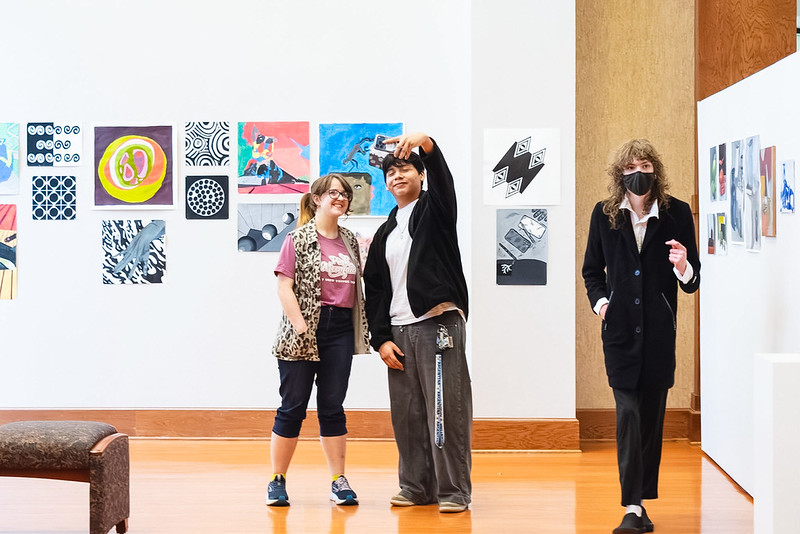 Three people in an art gallery with colorful abstract paintings; two smile for a selfie as a masked person in a coat walks by.