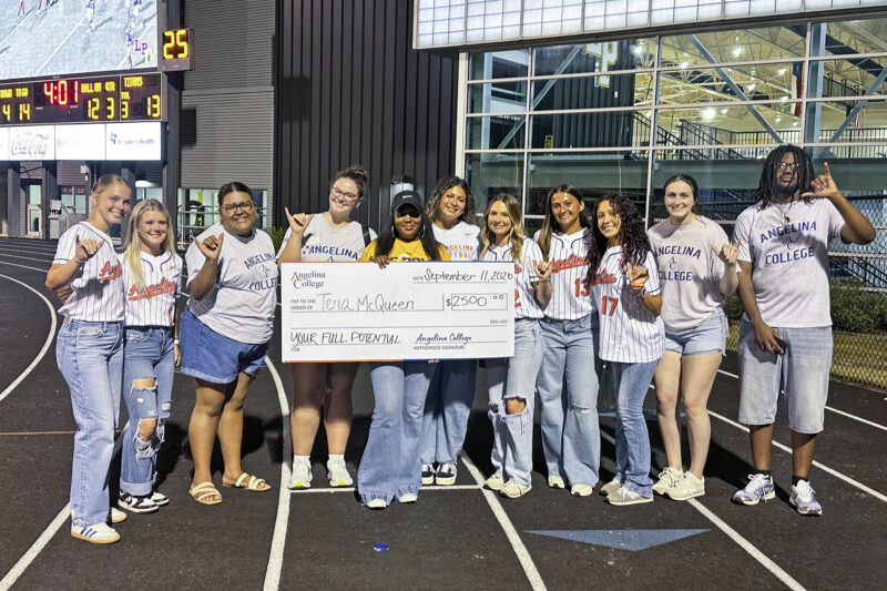 Smiling Angelina College students hold a $2,500 check for Tyna McQueen on a track at night, gym and scoreboard behind them.