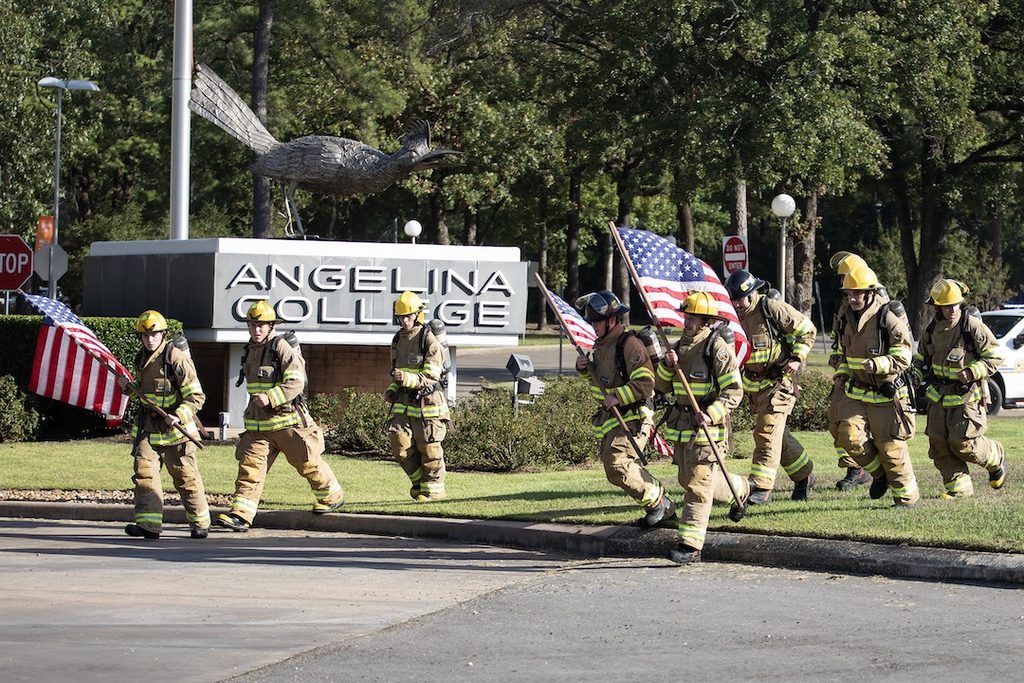 Firefighters in full gear carry American flags past a large Angelina College sign, with trees and a bird sculpture behind them.