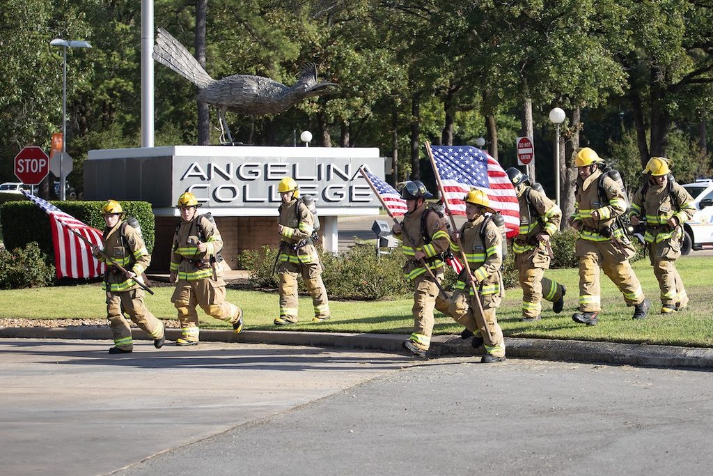 Firefighters in uniform walk by the Angelina College sign, carrying American flags with trees and a large bird statue behind them.