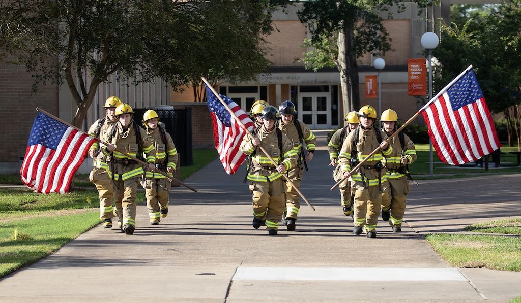 Firefighters in full gear carry American flags as they walk in formation on a path beside a building with trees and grass.