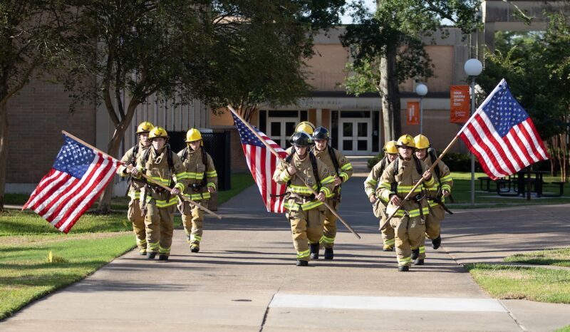 Six firefighters in uniform walk on a sunny sidewalk, carrying American flags. Trees and a brick building are in the background.