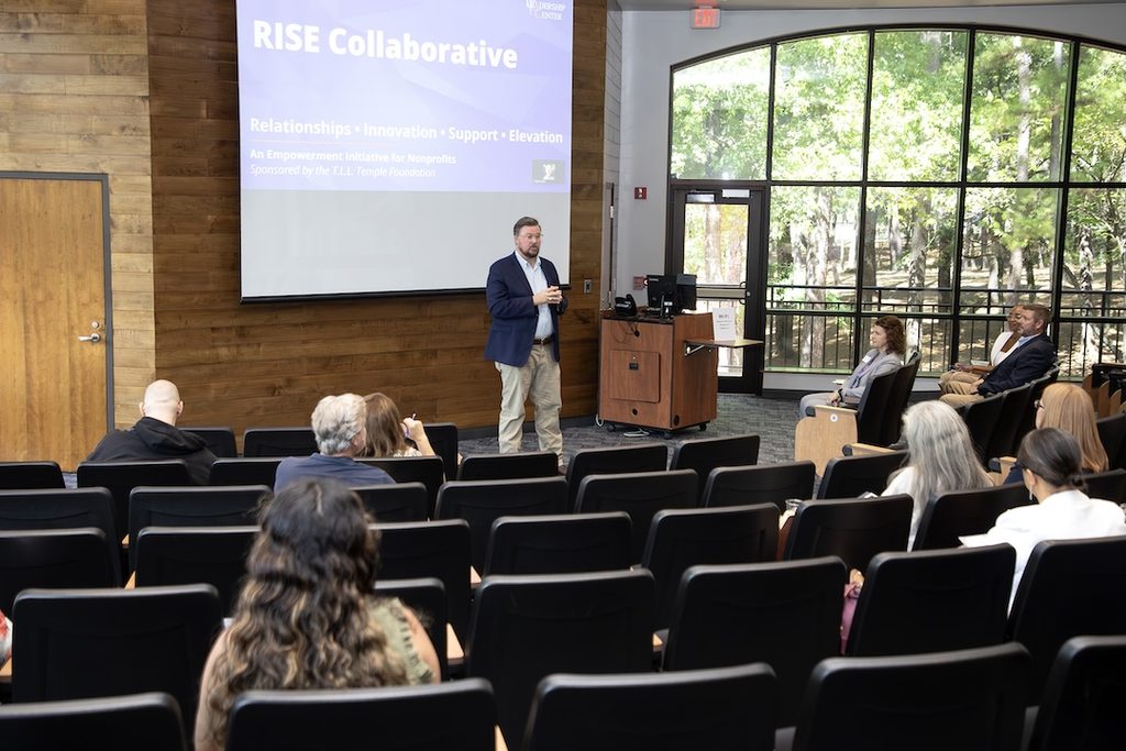 Man in a blue blazer speaks to a small audience; RISE Collaborative slide displayed behind him in an auditorium.