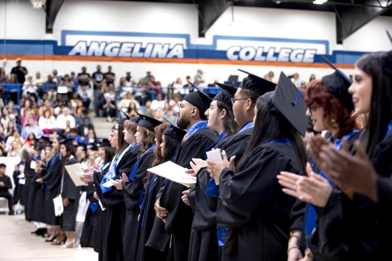 Graduates in caps and gowns line up at Angelina College as a crowd watches from gymnasium bleachers in the background.