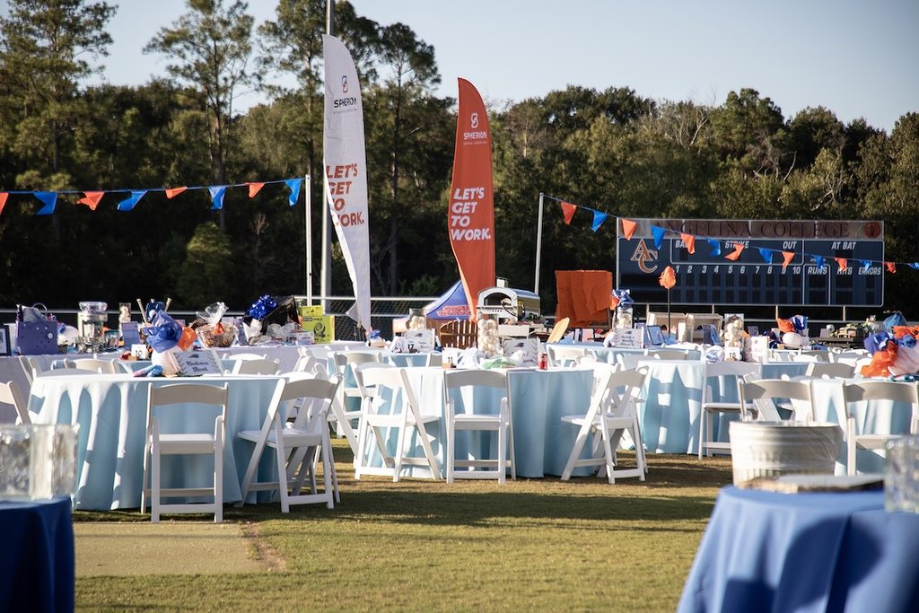 Round tables with blue cloths and white chairs arranged on a sports field; banners and a scoreboard in the background.