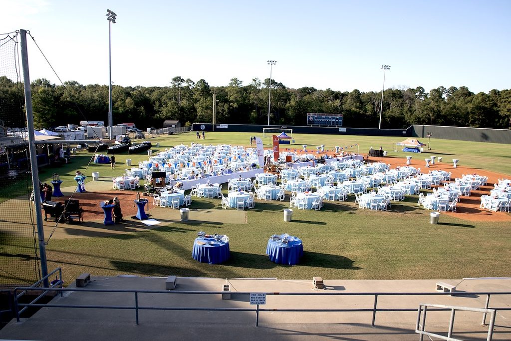 Round tables with blue and white cloths and chairs arranged on a baseball field under a clear sky for an outdoor event.