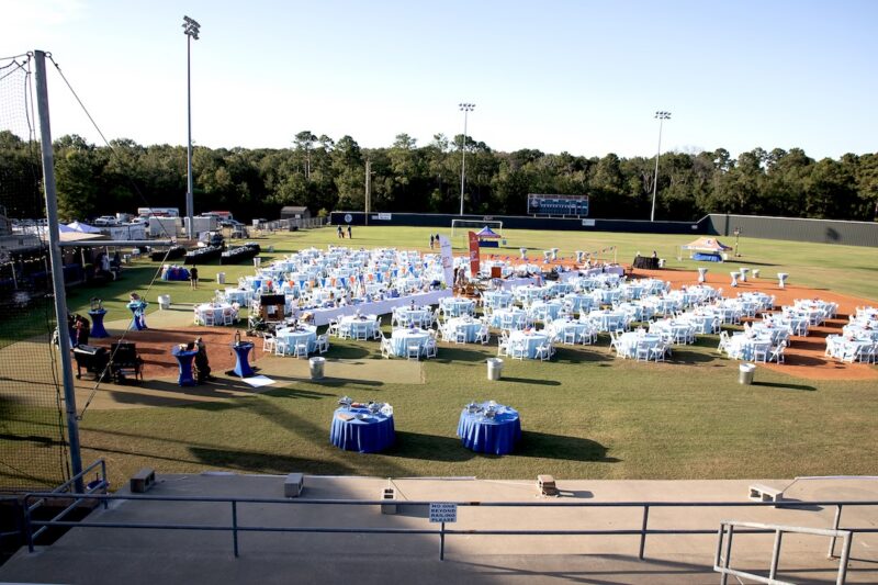 Outdoor baseball field with rows of white chairs and light blue tables, surrounded by greenery and sports gear.