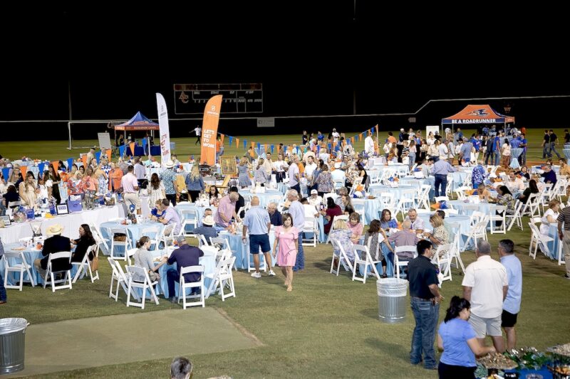 People enjoy an outdoor nighttime event with tables, orange and blue banners, tents, and lights on a grassy field.