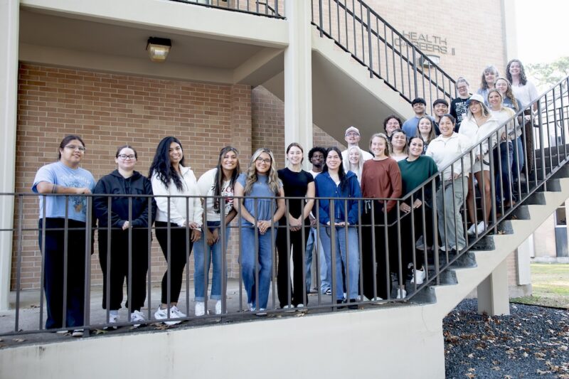 People of diverse ages and styles, casually dressed, smile at the camera while gathered on stairs outside a brick building.