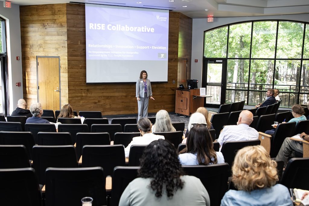 Woman speaks to an audience in a modern conference room; screen behind her reads “RISE Collaborative” with trees visible outside.