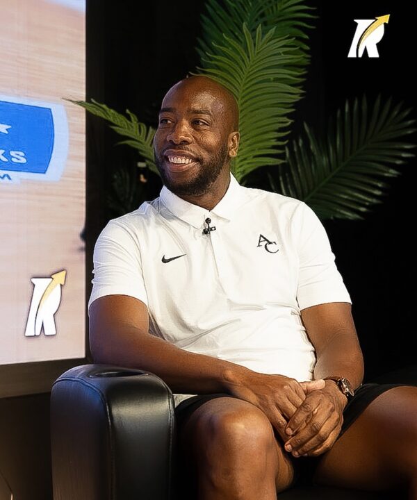Smiling man in white polo shirt sits in a black chair, hands folded, with green plants and a logo on a black wall behind him.