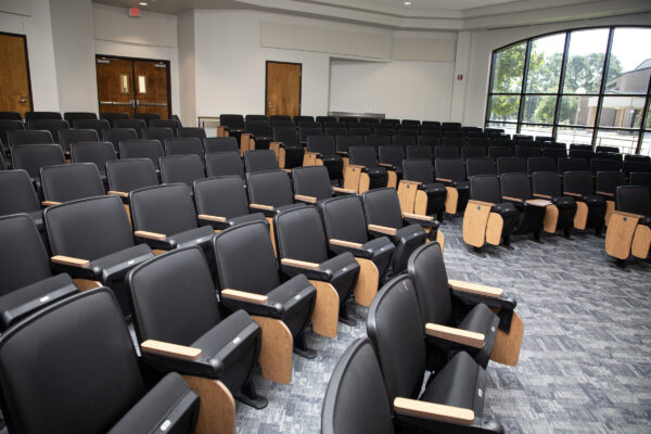 Lecture hall with empty black chairs, wooden armrests, grey carpet, large right-side windows, and sunlight streaming in.